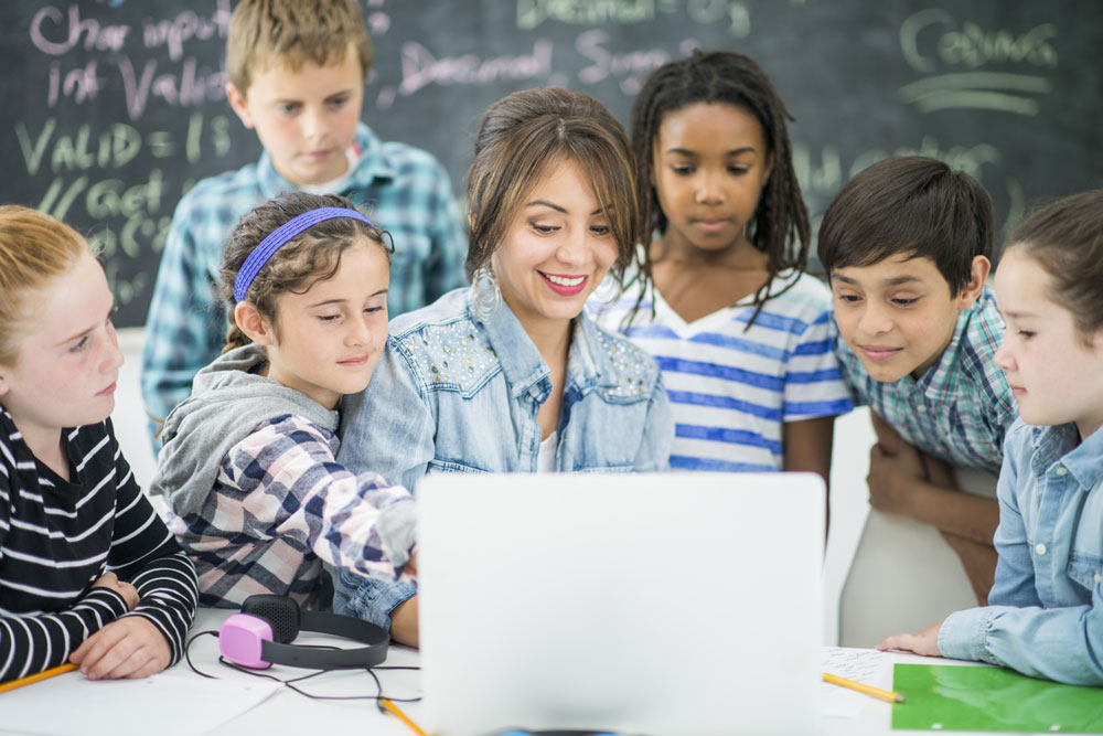 Young female teacher in a classroom surrounded by young children looking at laptop