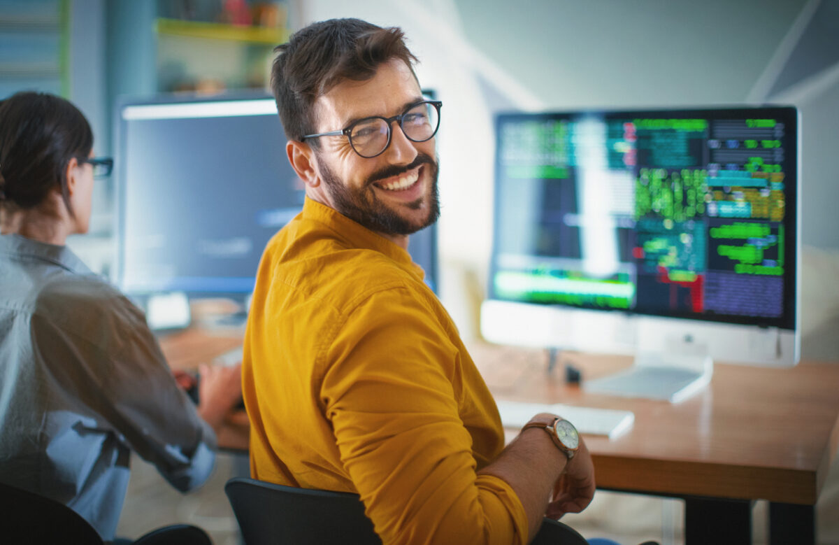 a man in a yellow jumper is leaning back in his chair and smiling over his shoulder. In front of him in blurred background is a computer screen.